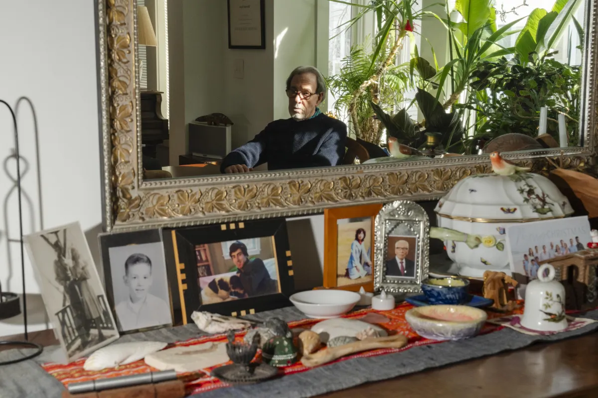 An older white man, seen in reflection in an ornate mirror, looks longingly as he's framed by personal knick knacks and photos adorning a table in front of said mirror