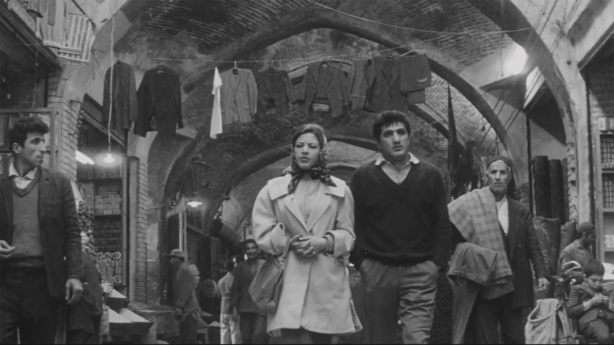 Black and white image of a woman in a coat and silk scarf over her hair walking alongside a man in pants ad a v-neck sweater around a Middle-Eastern market