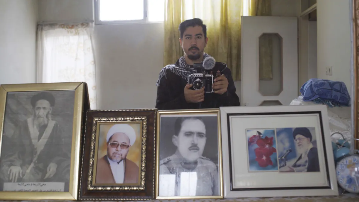A young brown-skinned man with short black hair and goatee beard holds a camera in front of a mirror where we can see various photos of older Middle-Eastern men in frames.