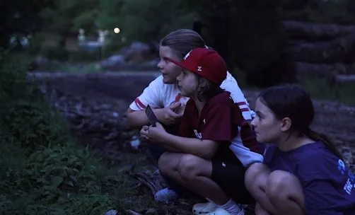 Three children at dusk in the woods crouched down looking into the trees and grass