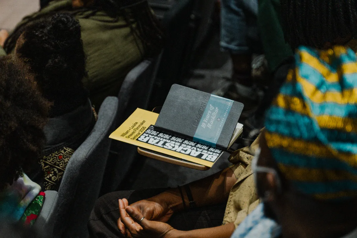 Close up shot of a Blackstar program and notebook on top of a desk, amid a number of Black folks seated as if for a lecture