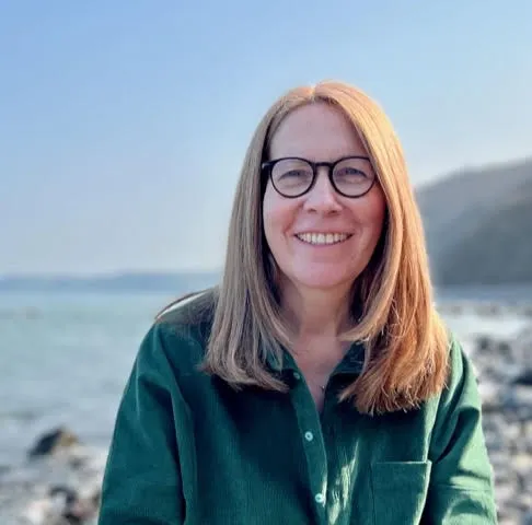 Headshot of a middle-aged white woman with shoulder-length auburn hair donning black-rimmed glasses smiling at the camera at a beach with a long-sleeve green button down shirt