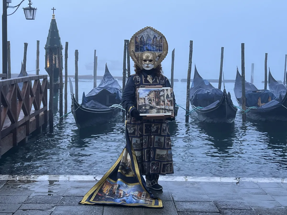 A masked figure in a Carnevale costume stands in front of gondolas and a dock at the edge of the island of Venice, Italy