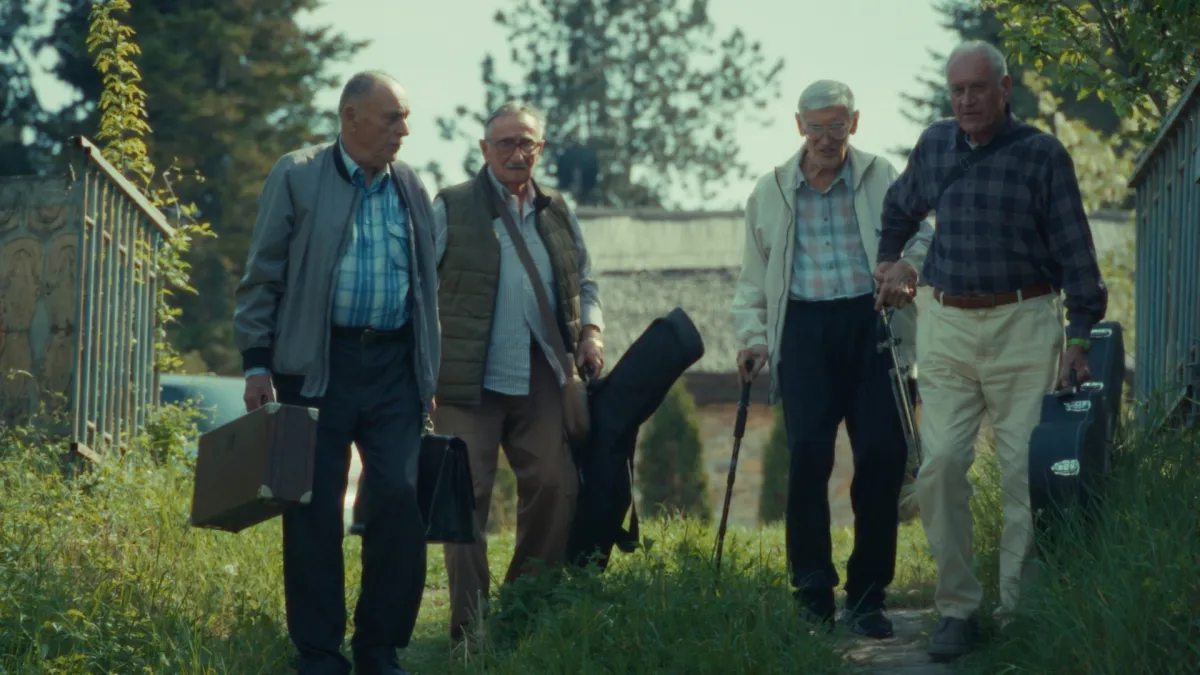 A group of elderly men in casual clothing carrying instruments in cases walk alongside a countryside road