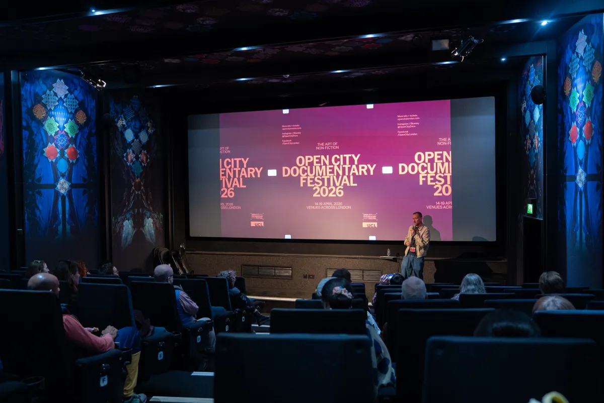A white man stands in front of a crowded theater, a screen behind him showcasing an ad for the Open City Documentary Festival