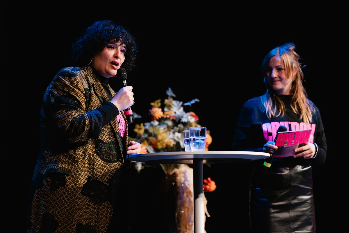 A light-skinned woman with shoulder-length curly black hair in a stylish printed coat holds a mic at a table with two glasses as a young blond-haired woman wearing a CPH:DOX tee stands in the background