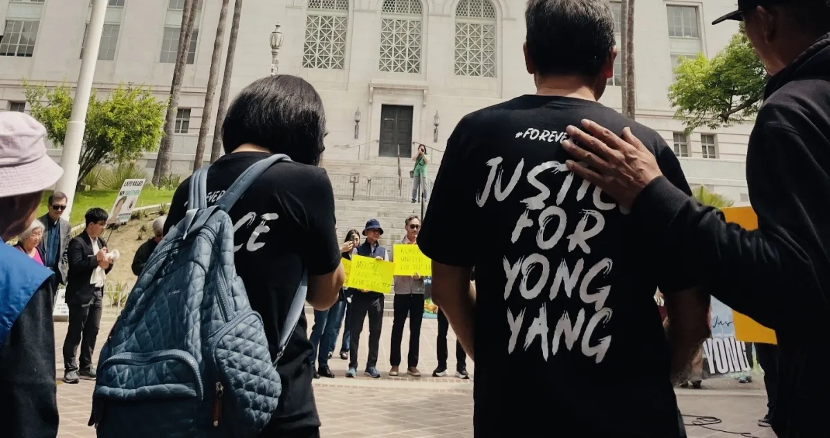 A peaceful outdoor rally taking place on the steps of Los Angeles City Hall. In the foreground, two people are seen from behind, wearing black t-shirts that read "Justice for Yong Yang." Behind them, a diverse crowd of participants of various ages, some wearing hats and sunglasses, hold signs that read "Mental Health Lives Matter" and "Koreans United for Justice."