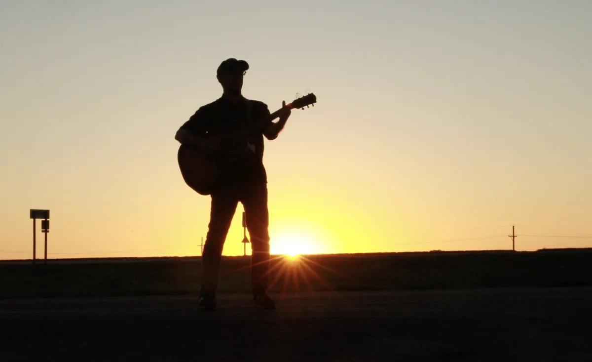 A lone man plays an acoustic guitar on the side of a freeway, silhouetted by the sunset at his back
