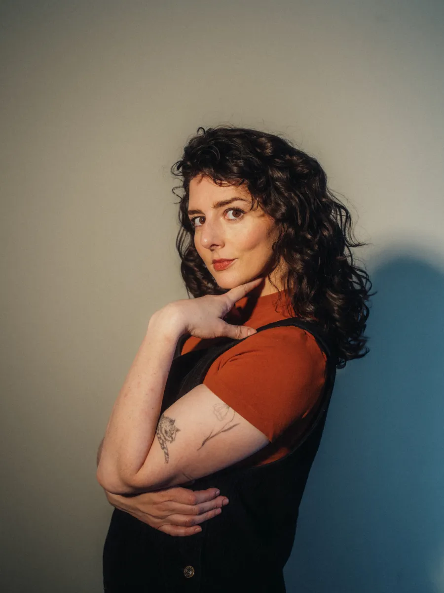 Photo of a fair-skinned woman with dark brown hair. She is wearing a red shirt under a black smock. 