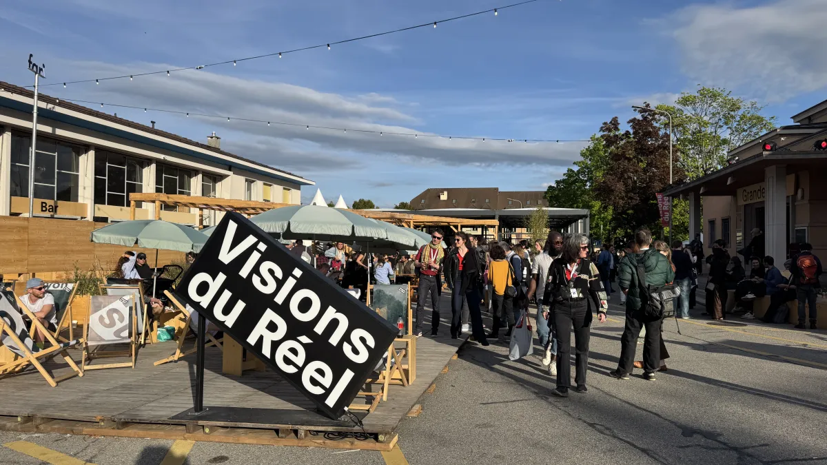 Under a crisp blue sky, a crowd of festivalgoers stands under the outdoor umbrellas. In front of them is a large sign that reads: "Visions du Reel."