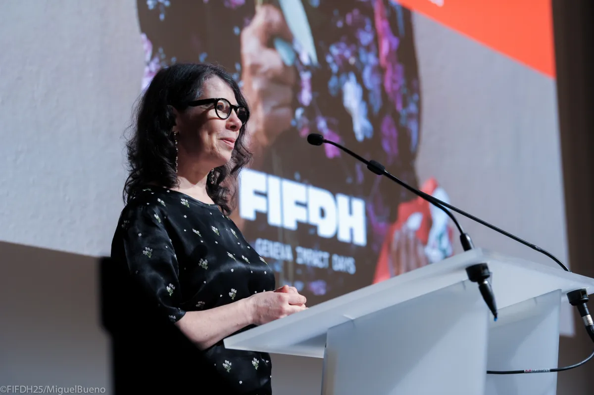A middle-aged white woman with shoulder-length black wavy hair dons glasses and a black dress with white flowers at a podium with a mic