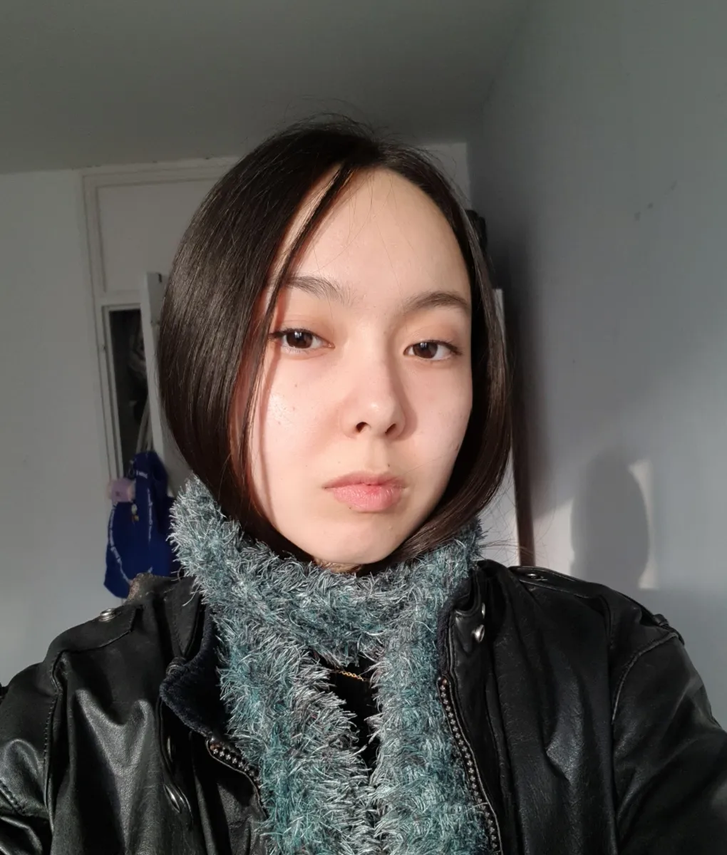 Headshot of a young Asian woman with long black hair tucked into a blue-ish scarf and black jacket poses for the camera