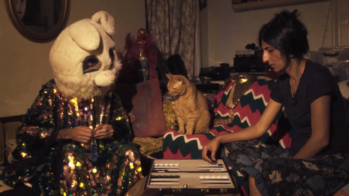 A figure in a colorful, sequined robe and bunny mask and a woman in a black shirt play backgammon. An orange cat sits between them, appearing to stare incredulously at the masked figure