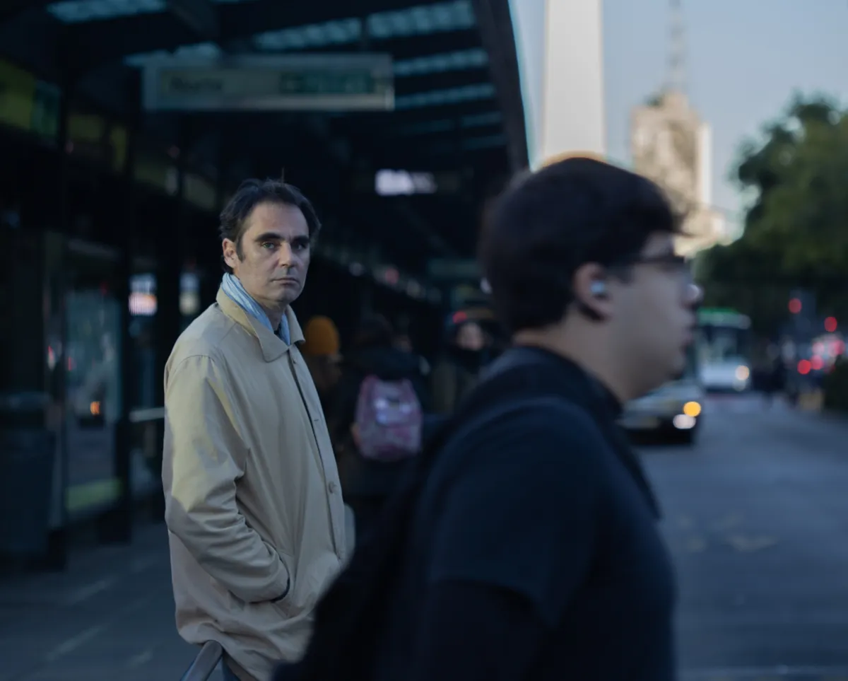 Middle-aged white man in a khaki rain coat over a light blue shirt stands in a somewhat crowded street looking straight at the camera