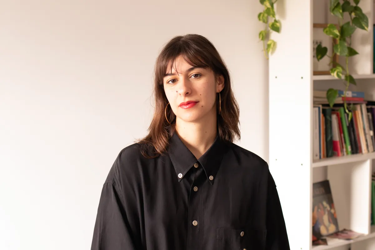 A young white woman with shoulder-length dark brown hair with bright red lipstick stares at the camera in an oversized black shirt against a mostly blank wall behind her