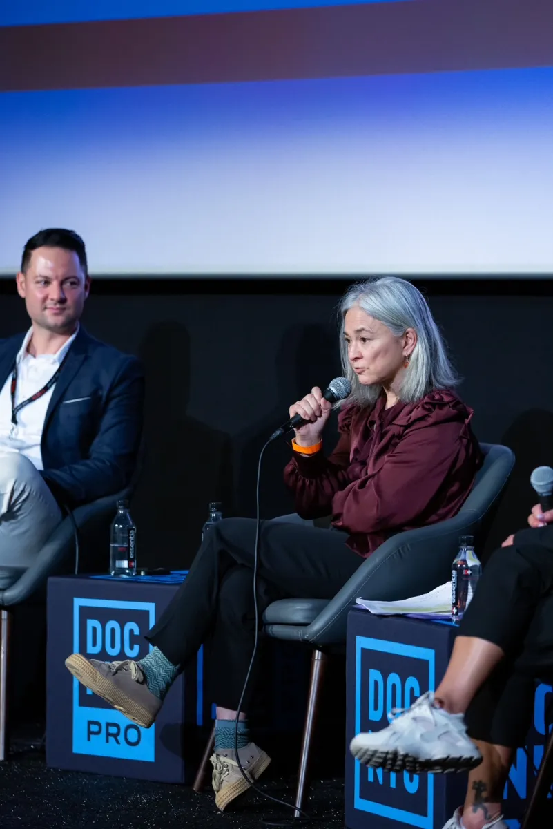 A woman in a maroon button-down shirt speaks into a microphone. She sits on a chair infront of a large screen. A man in a white shirt and blue blazer sits to her right. Both face forward. Tables on either side and in-between the two read "DOC NYC PRO"