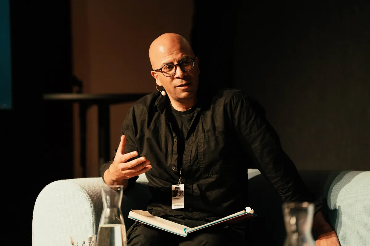 Bald man in glasses and dark brown collared shirt sits in white arm chair, gesturing with hand