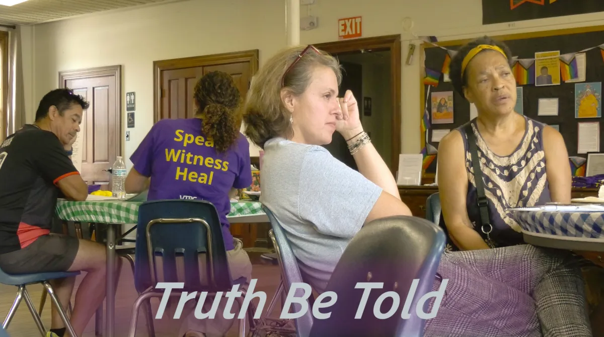 A man on the left in a black shirt at a table with a woman who is wearing a purple shirt with the words on the back: Speak, Witness, Heal.  A woman featured in the center of the photo is leaning back in her chair, legs crossed with her hand on her head looking off camera.  A black woman with a gold hair band is sitting next to the woman looking off camera.