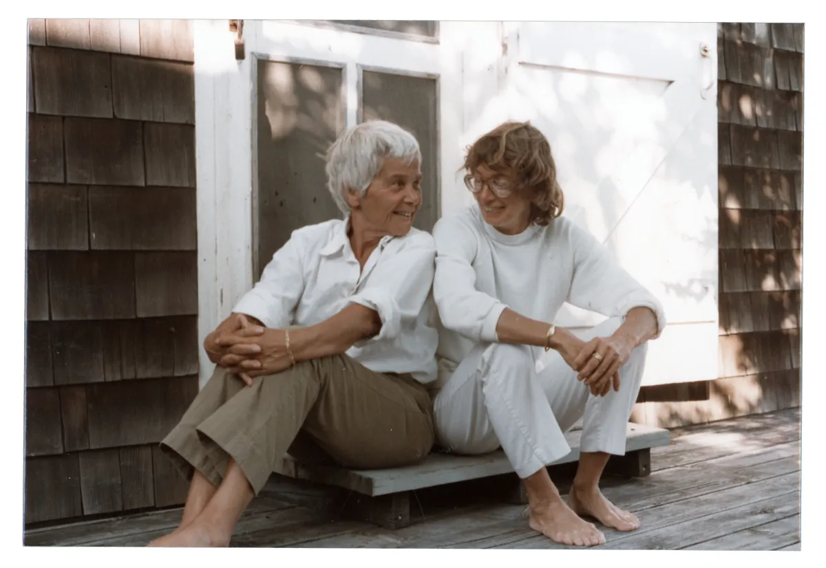 Two women sit on a doorstep smiling at each other. On the left is an older woman with short white hair in khaki pants and a white collared shirt. On the right is a woman with mid-length brown hair, glasses, a white turtleneck and white pants. Both are barefoot