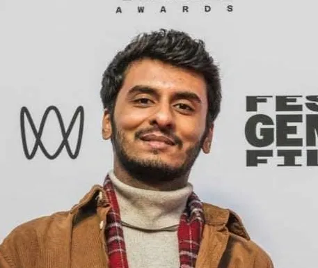 Headshot of a smiling man with short dark hair and facial hair standing in front of festival photo backdrop. He wears a white turtleneck, red plaid scard, and yellow collared shirt