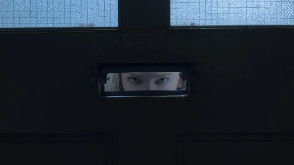 A blonde woman's blue eyes can be seen through a slot in an otherwise black surface with two windows visible at the top of the frame