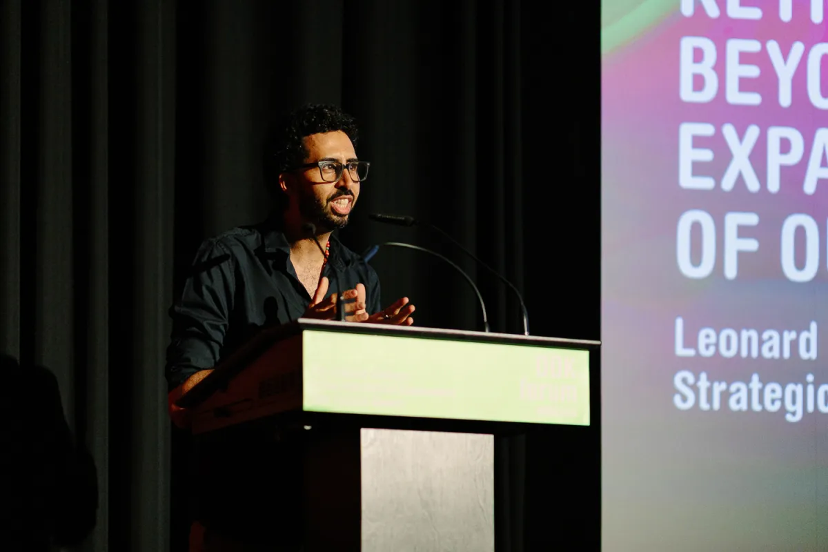 A man in a black shirt with black glasses and short dark hair and beard delivers a speech at a podium. Behind him is a black curtain and the lefthand side of a pink and green digital display showing text which cannot be read