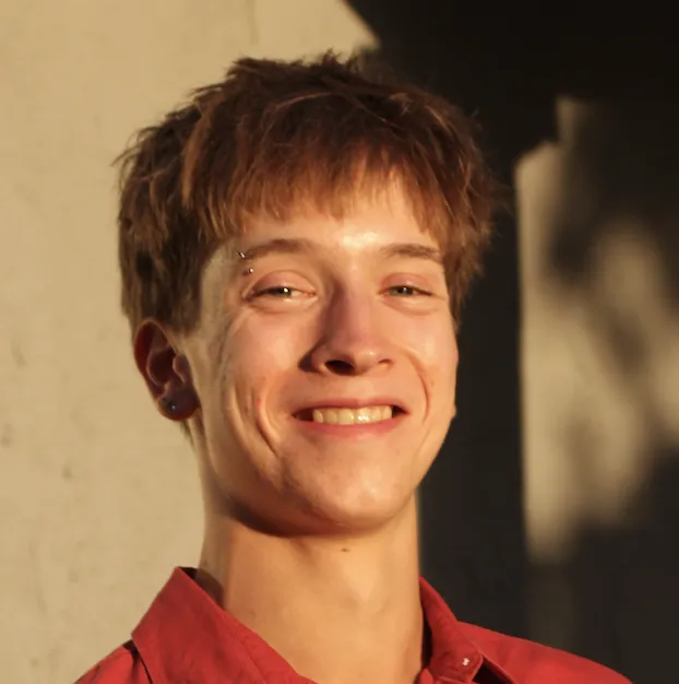 Headshot of a 21 year old white man with short blond hair and a pierced eyebrow, wearing a red collared shirt and smiling
