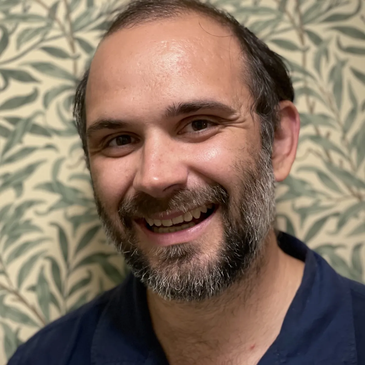 Headshot of a smiling man with short dark hair and greying facial hair in front of floral wallpaper
