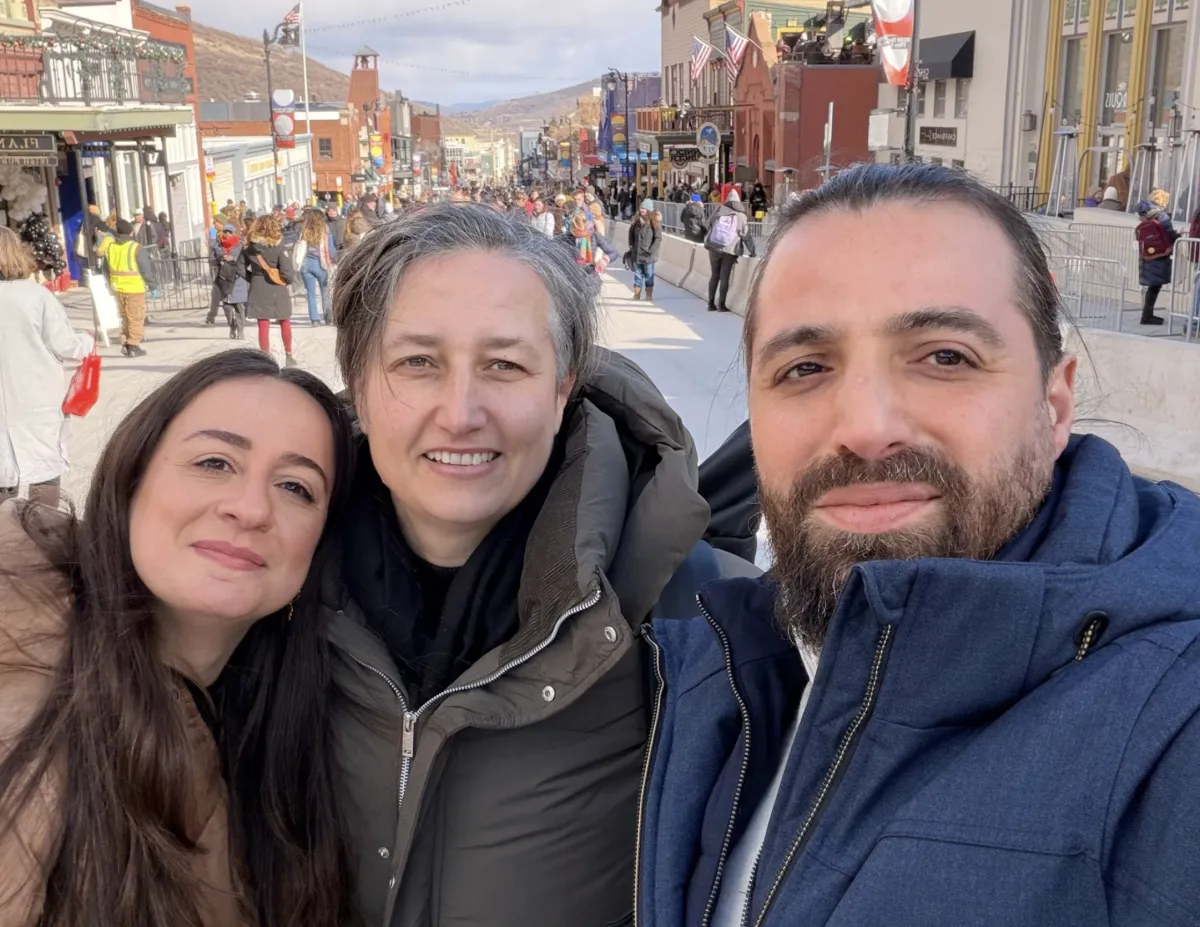 Selfie taken by a bearded man (right), woman with short grey hair (center), and woman with long, dark hair (left). All wear warm coats and smile. The background is a street lined with shops and pedestrians