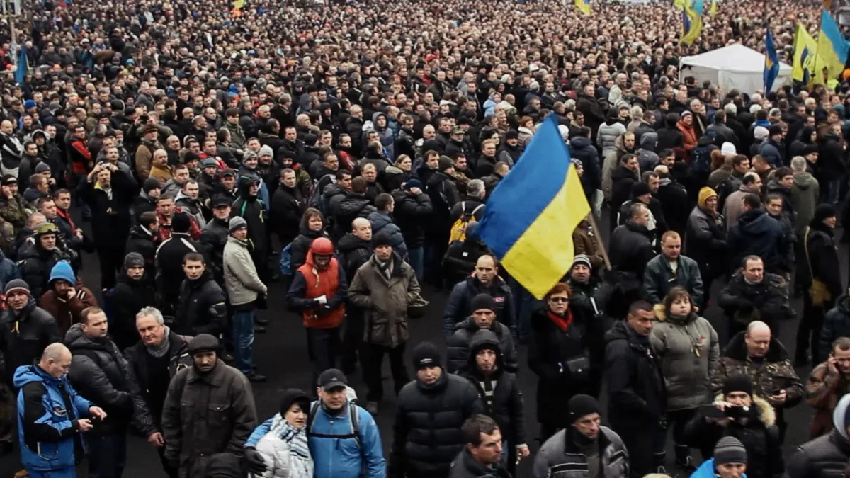 A massive crowd of people in winter clothes is gathered. One man towards the front waves a large Ukrainian flag