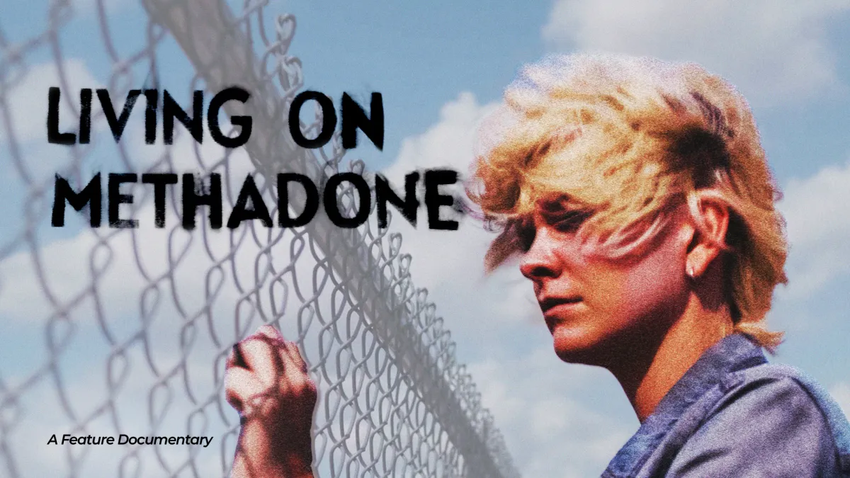 The image shows a young person with short, windblown blond hair standing beside a chain-link fence under a bright blue sky with large white clouds. They are wearing a gray denim-style jacket and appear to be looking through or past the fence with a quiet, reflective expression.  The composition feels cinematic and symbolic. The fence creates a strong visual barrier, suggesting themes of separation, confinement, longing, or being caught between two worlds. The open sky behind them adds contrast, giving the i
