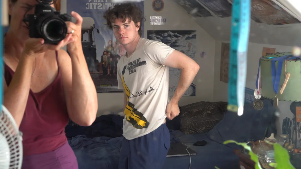 A young white man with short curly dark hair in an Andy Warhol white tee stares at a mirror in his bedroom as a white woman whose face is obscured by a camera takes a photo of them both
