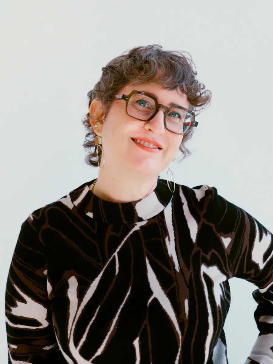 A woman with fair skin smiles at the camera. She has short, curly hair, green eyes, and is wearing a patterned shirt. 