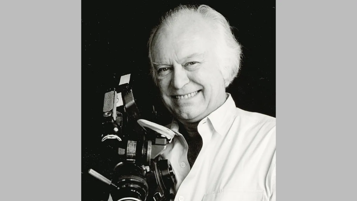 Black and white headshot of an older white balding man with tufts of white hair who smiles at the camera while holding a film camera