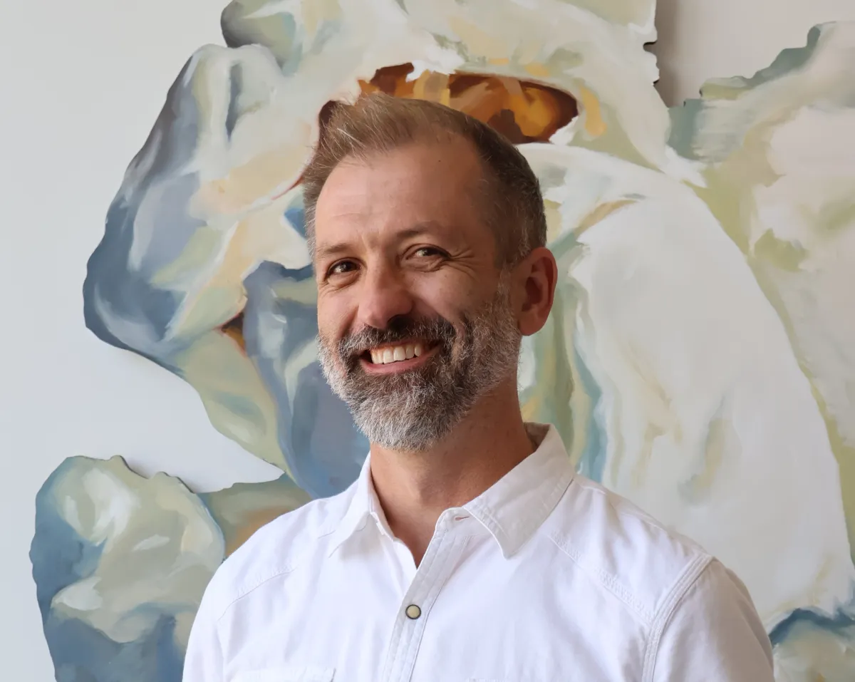 A smiling white man in a white shirt with thinning light hair and a salt and pepper beard stands in front of a striking piece of art in muted colors