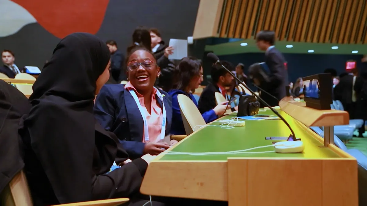A candid photograph inside the United Nations General Assembly Hall captures two young delegates sharing a joyful moment between sessions. One student, wearing a black hijab, sits facing another delegate in a suit and conference lanyard, who is laughing openly. Around them, other students talk, move between seats, and prepare for proceedings, with microphones and the iconic General Assembly desks visible throughout the frame.  The image captures the heart of The Delegates: young people participating in seri