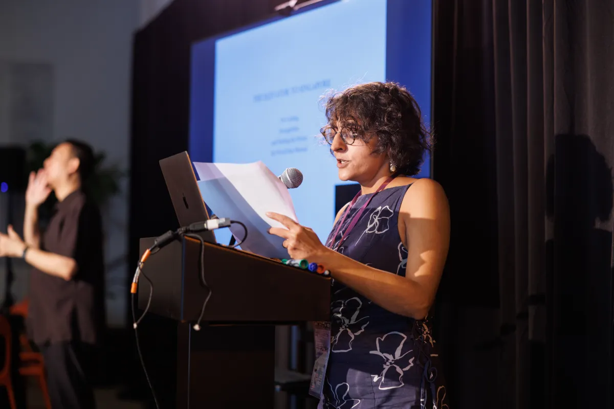 A woman (Sarvnik Kaur) with short, curly hair in a floral dress and glasses stands at a podium, speaking into a microphone. Behind her, out of focus, are a sign language interpreter and a large screen displaying text on a white background.