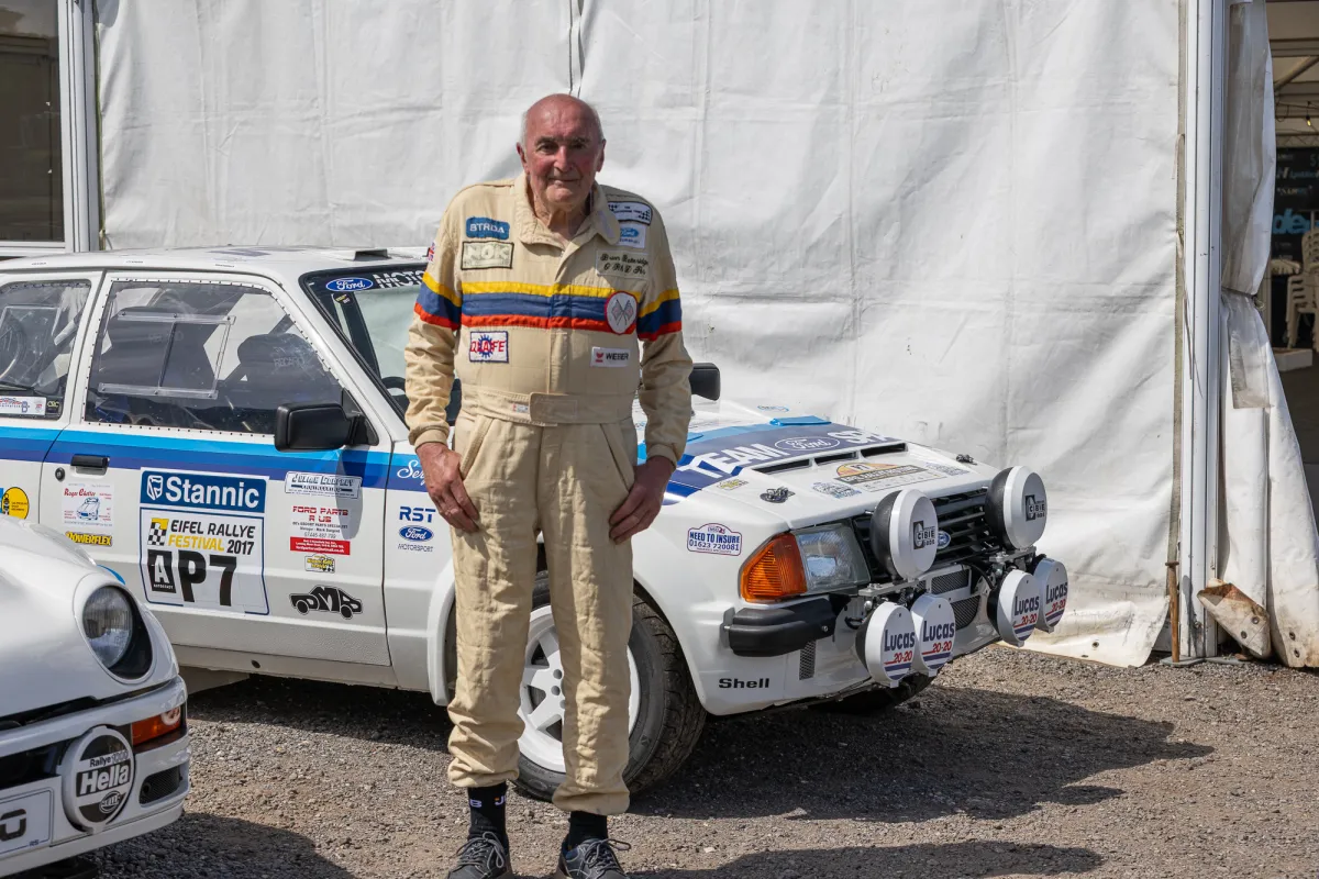 Brian Betteridge wearing a cream racing suit stands in front of his white Ford Escort RS1700T rally car with blue stripes and sponsor decals.