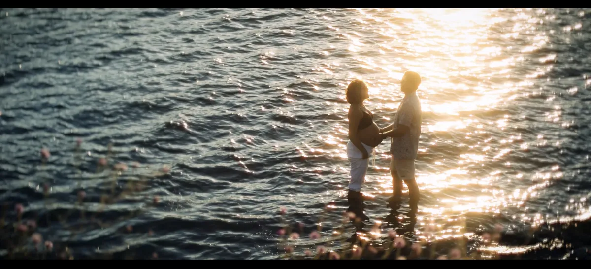 A beautiful, serene scene of a man and woman standing in the water at sunset as the man holds the woman's pregnant belly.