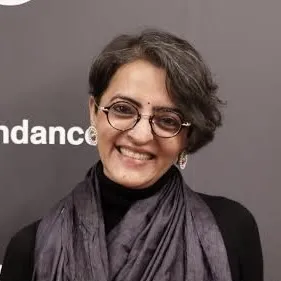 A smiling woman in a silk scarf, black turtleneck, and glasses with a short side part stands in front of a background which reads 'sundance'