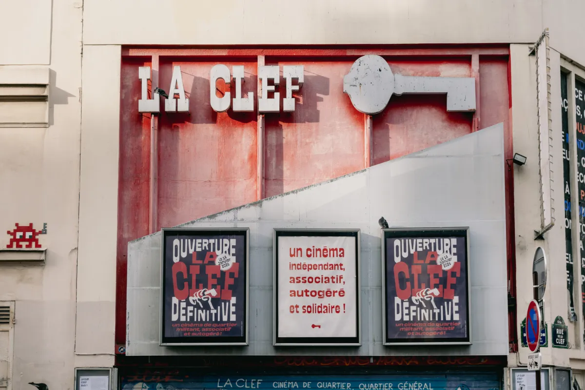 The front of a building, half red and half grey. At the top are the words "LA CLEF" and the shape of a key in grey. Three large posters are displayed on the building, reading "Ouverture La Clef Dēfinitive" and and "Un cinēma indēpendant, associatif, autogērē et solidaire!"