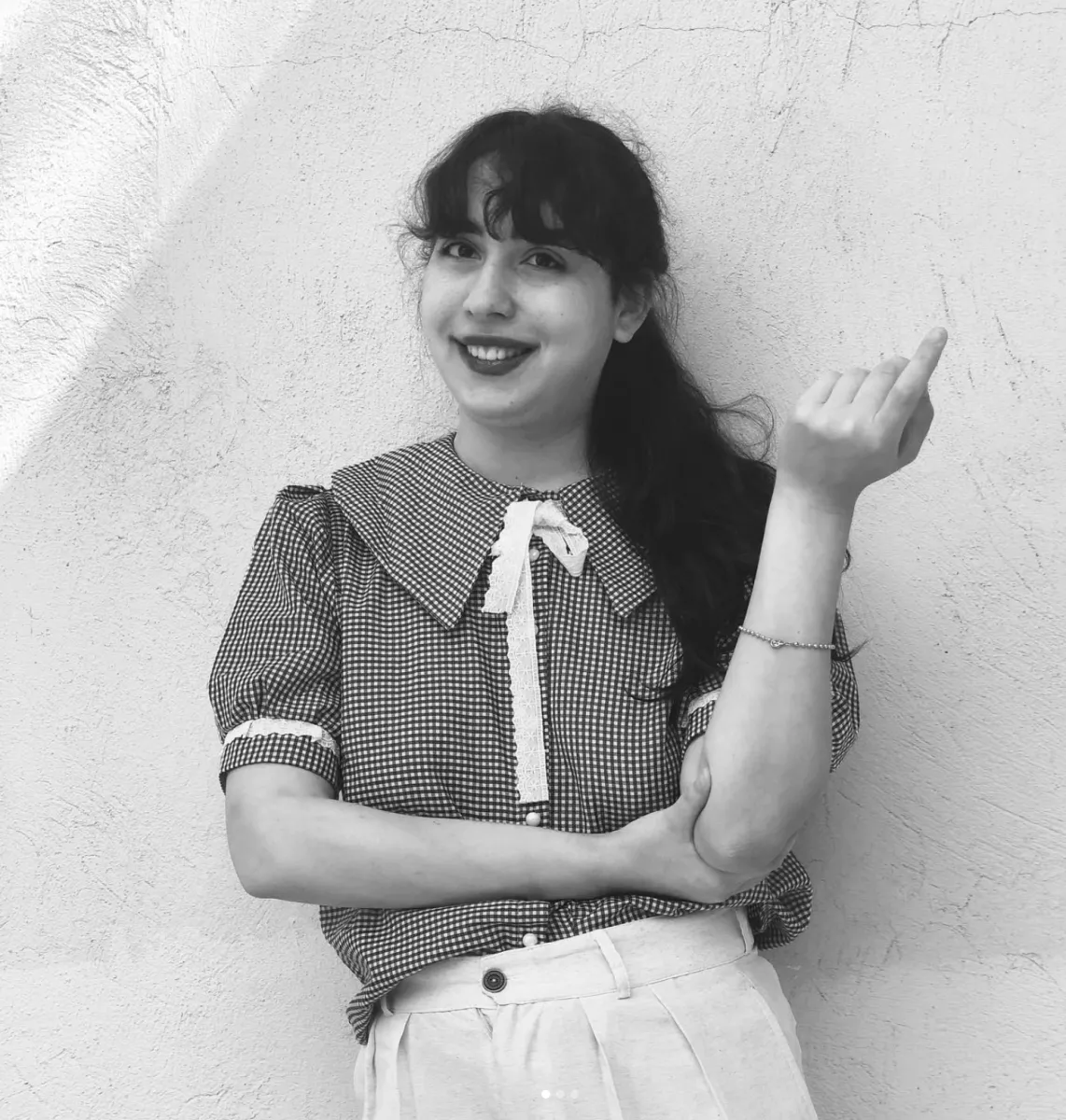 Black and white portrait of a smiling woman with long, dark hair leaning against a wall wearing a plaid shirt with a large collar and pleated white pants