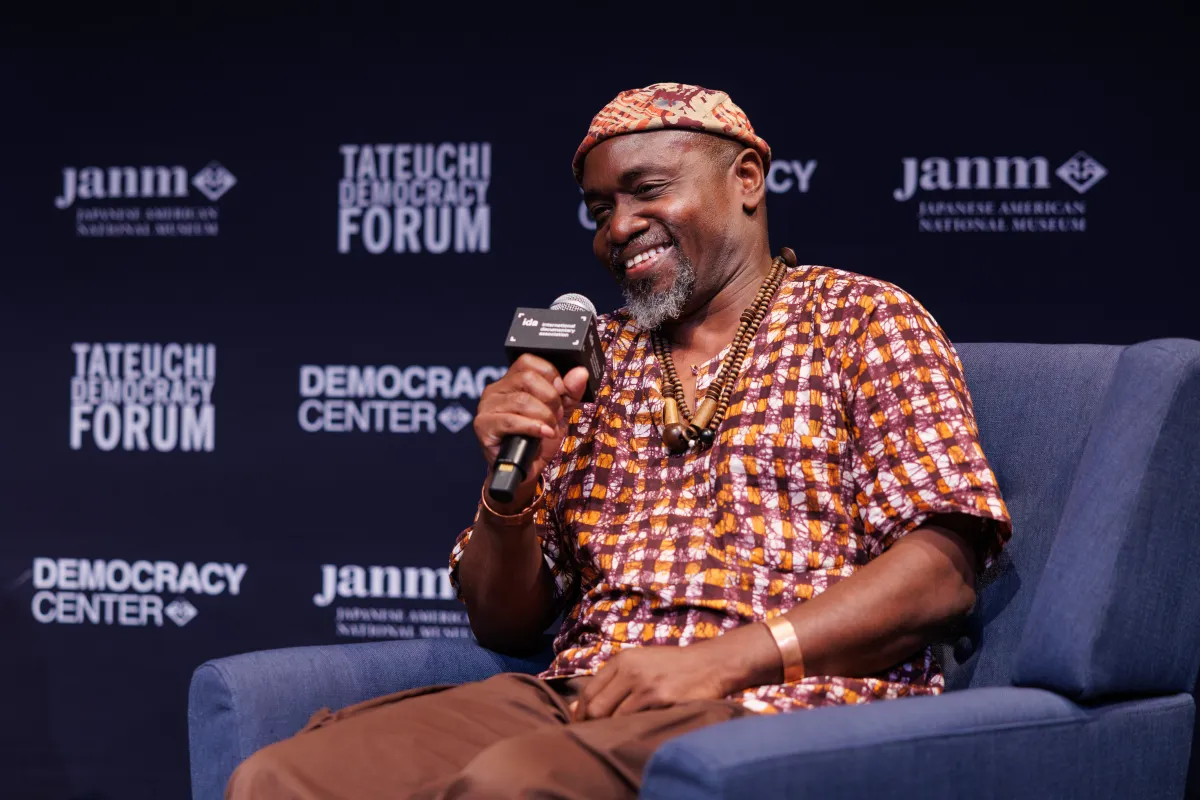 A smiling man in a shirt and hat composed of the same orange, brown, and white patterned fabric, wearing beads around his neck and a metal bracelet, holds a microphone labelled "ida international documentary association" and sits in a blue armchair. Behind him is a black background with white text reading "TATEUCHI DEMOCRACY FORUM", "janm", and "DEMOCRACY CENTER"