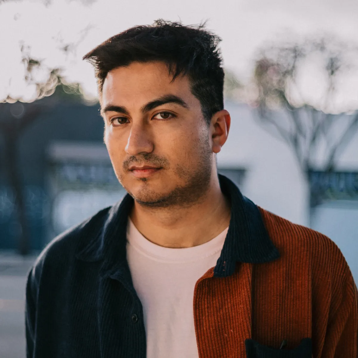 Headshot of a young man with short, dark hair, wearing a rust-colored collared shirt over a white T-shirt. The background is a neighborhood out of focus