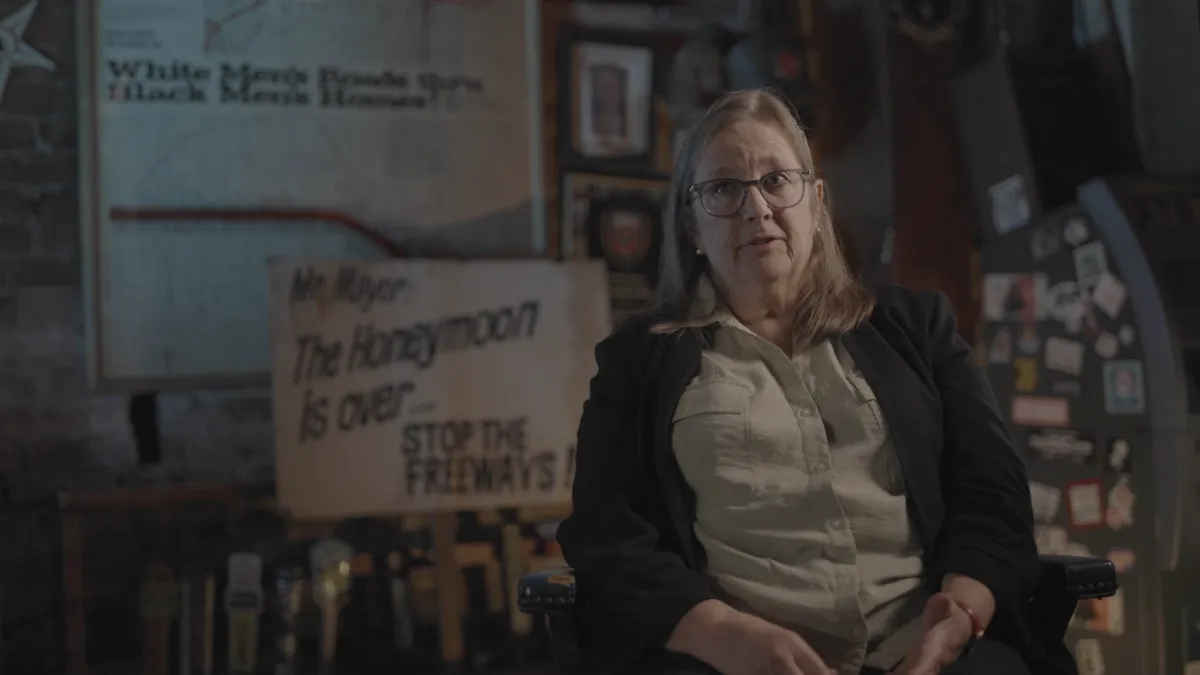Image shows Kate Kelly, an older woman and resident of the brookland neighborhood who was a witness to the protests at the time. She is sitting being interviewed with a poster and signage from the protests blurred but still visible behind her. They say; "Mr. Mayor the honeymoon is over...stop the freeways!" and "White men's roads through black men's homes!"