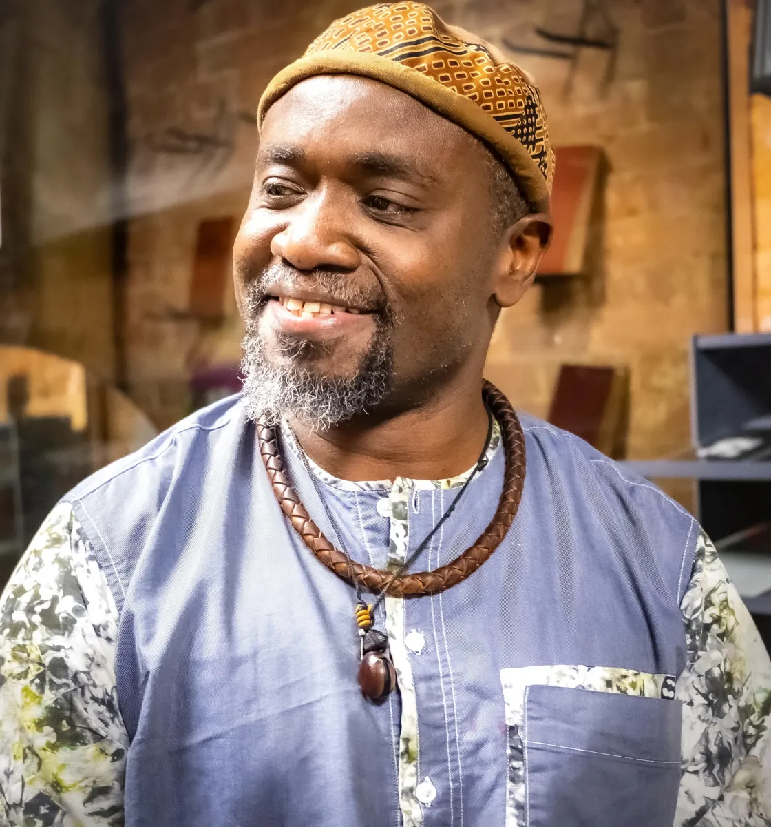 Headshot of a smiling Black man with a short beard, a yellow cap, a blue jacket with white and green sleeves and accents, and two necklaces, in front of a background that appears to be backstage of a theater