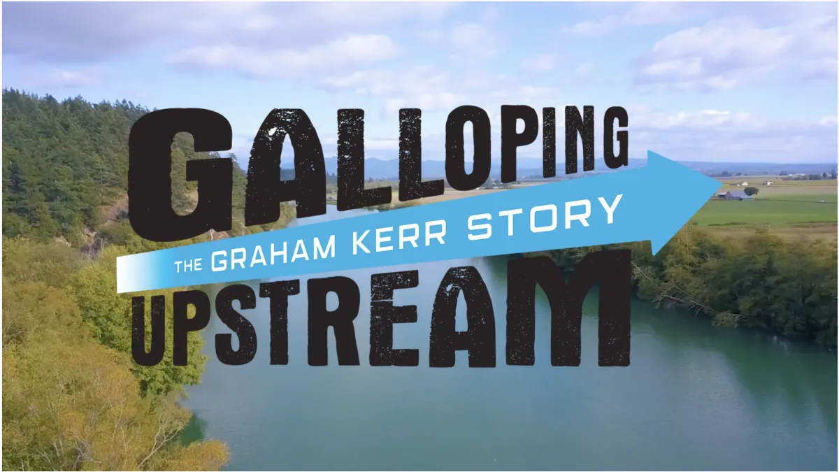 A cinematic aerial view of a winding river surrounded by lush green forests and open farmland under a blue, cloud-filled sky. Bold, distressed black lettering reads “GALLOPING UPSTREAM,” crossed by a bright turquoise arrow containing the white subtitle “THE GRAHAM KERR STORY.” The design feels adventurous, energetic, and inspirational, suggesting a journey of perseverance and discovery.