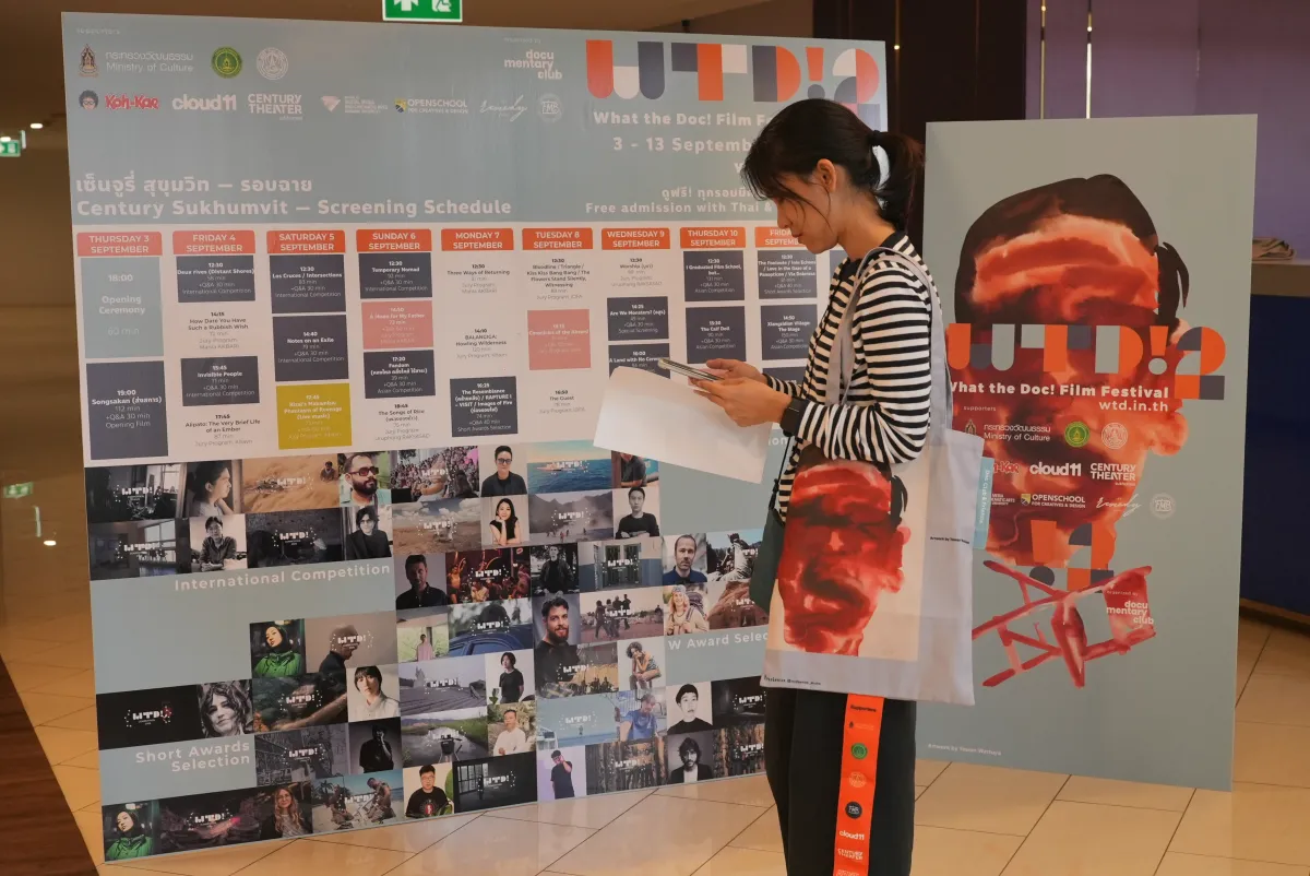 A woman in a striped shirt with a tote bag over her shoulder uses her smartphone. She stands in front of two large banners, one of which displays a schedule for the 2026 edition of What the Doc!, and one of which displays a graphic of a man's face (the same graphic which is on her bag) behind the words What the Doc! 2