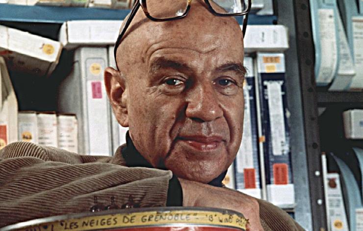 A middle-aged Bud Greenspan posing in front of film tapes. He is bald and is wearing black framed glasses on his head.