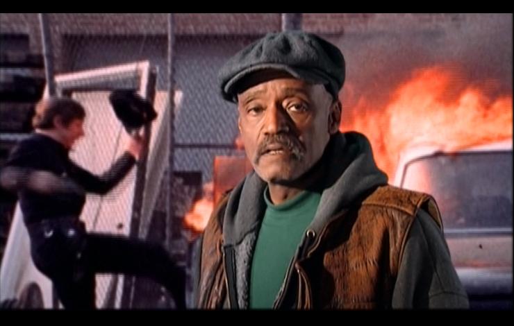 A Black man stands in front of an inflamed car, from Melvin Van Peebles' 'Classified X.' 